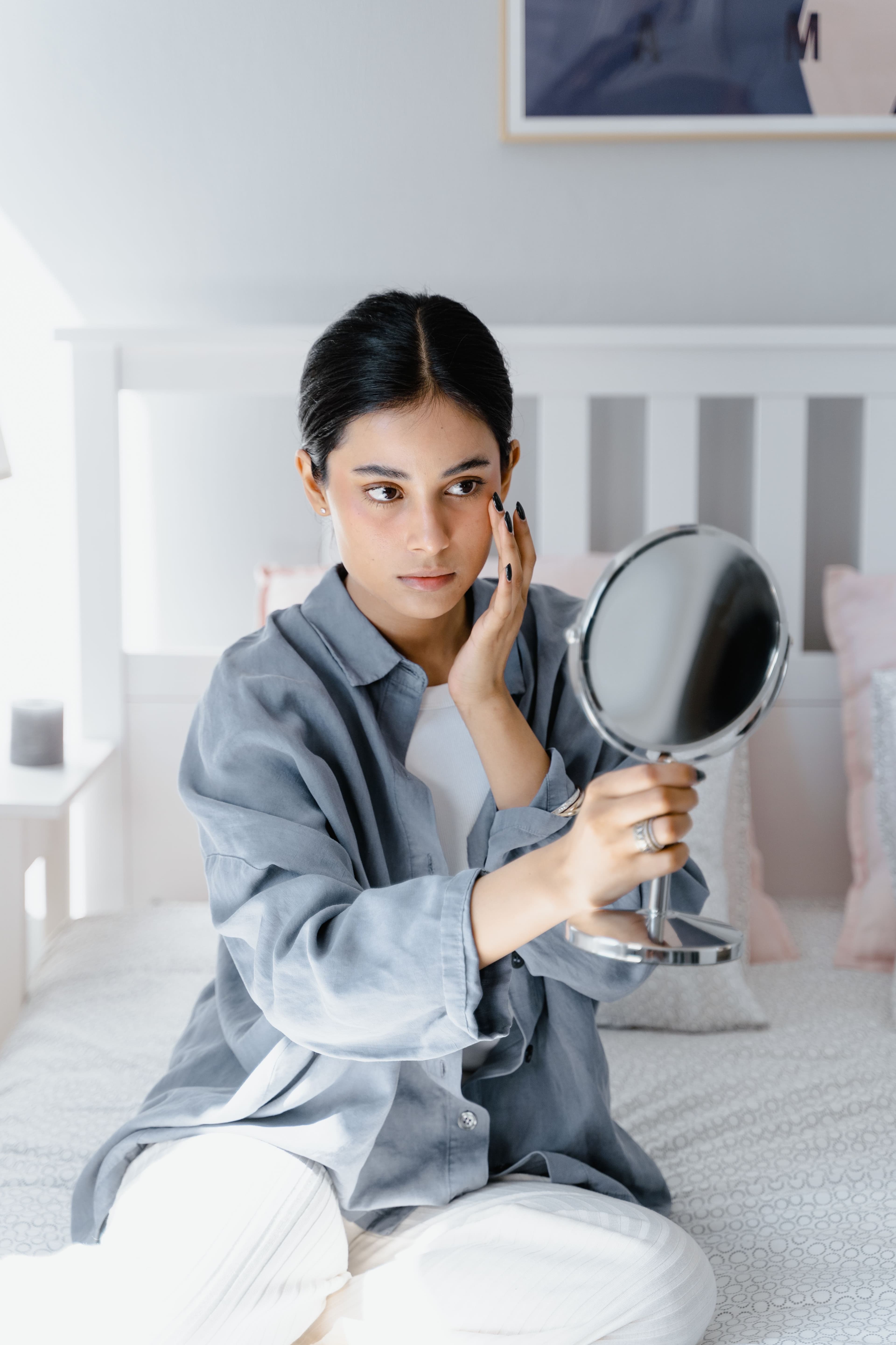 Woman sitting on bed analyzing her face in handheld mirror.