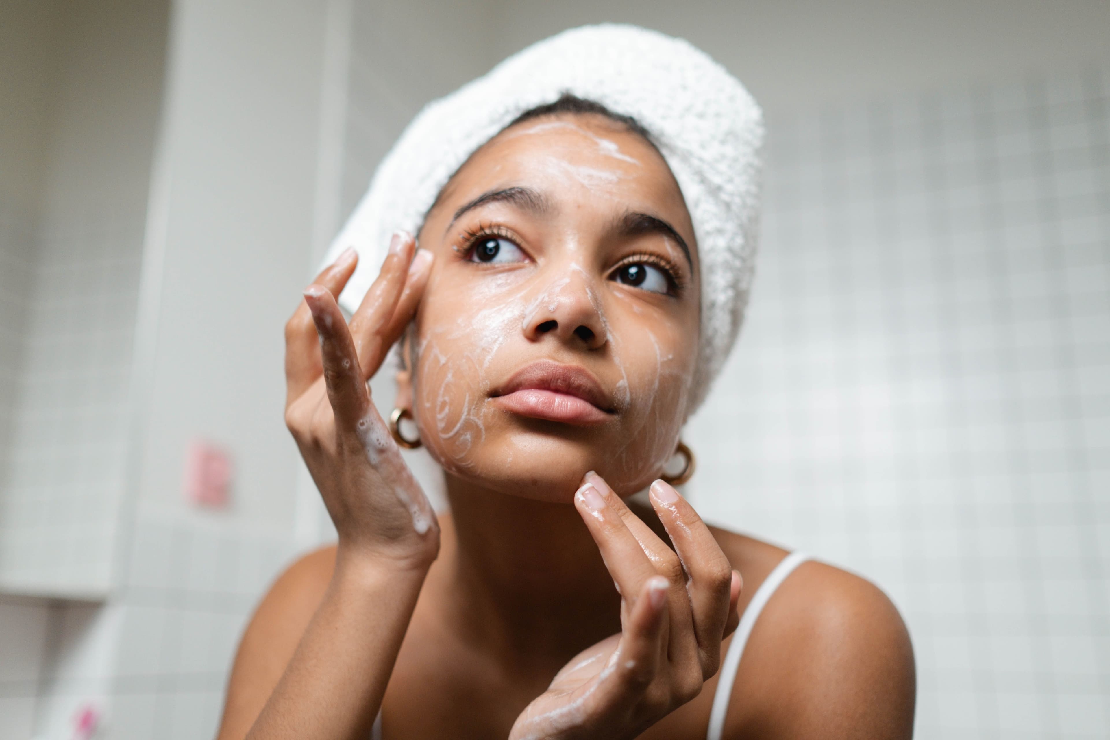 Woman with towel on her head is looking at herself in mirror while applying cream to her face,